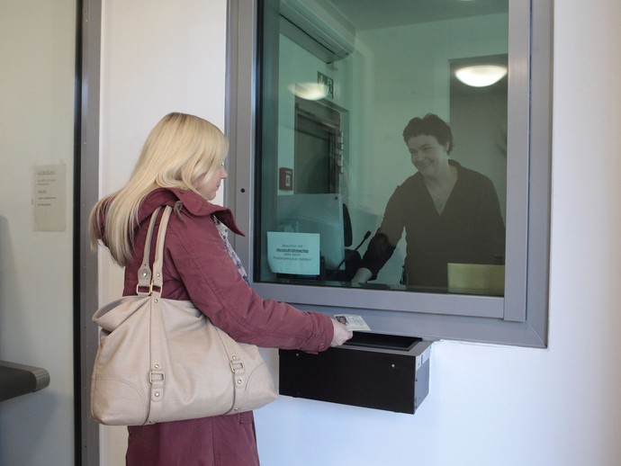 Eine Frau steht hinter einer Scheibe und nimmt den Ausweis einer Besucherin entgegen. (Foto: LWL)