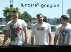 Drei junge Männer laufen lachend auf eine gläserne Tür mit der Aufschrift "Personaleingang" zu