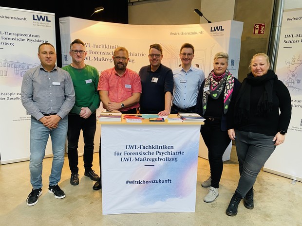 Fünf Männer und zwei Frauen stehen an einem Infostand mit der Aufschrift "LWL-Maßregelvollzug" und lächeln in die Kamera (Foto: LWL/Hekel) Fünf Männer und zwei Frauen stehen an einem Infostand mit der Aufschrift "LWL-Maßregelvollzug" und lächeln in die Kamera (Foto: LWL/Hekel)