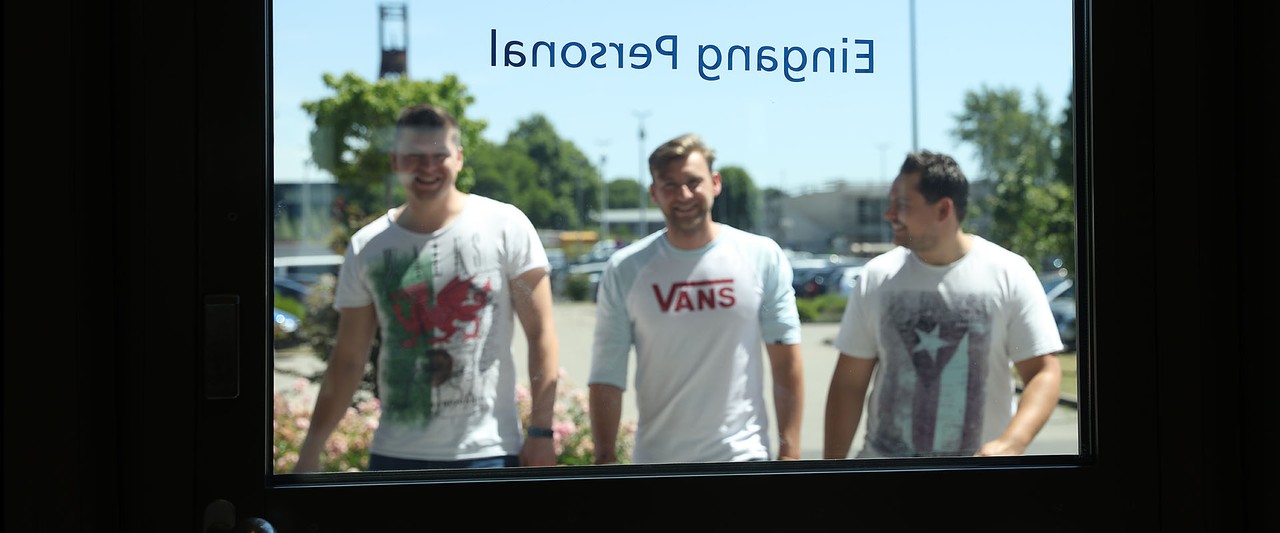 Blick auf eine Glastür, auf die von außen drei junge Männer zulaufen
