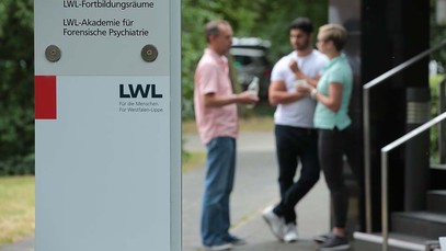 Im Vordergrund ein Schild mit der Aufschrift "LWL-Akademie für Forensische Psychiatrie", im Hintergrund drei plaudernde Personen