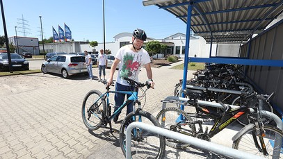 Ein Mann schiebt sein Fahrrad in den überdachten Fahrradständer, während im Hintergrund zwei Männer auf dem Klinikparkplatz warten.